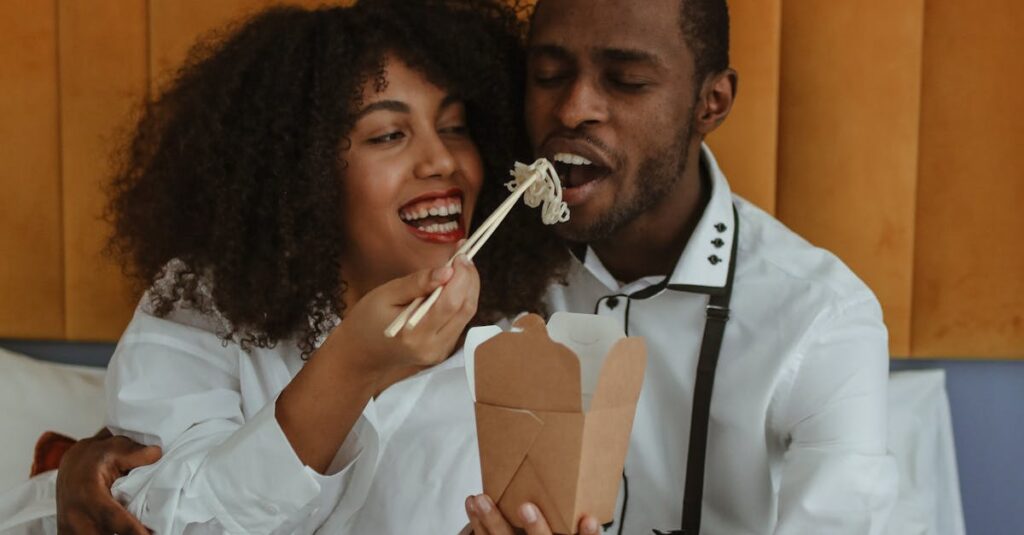 Smiling couple sharing noodles from a takeout box using chopsticks indoors.